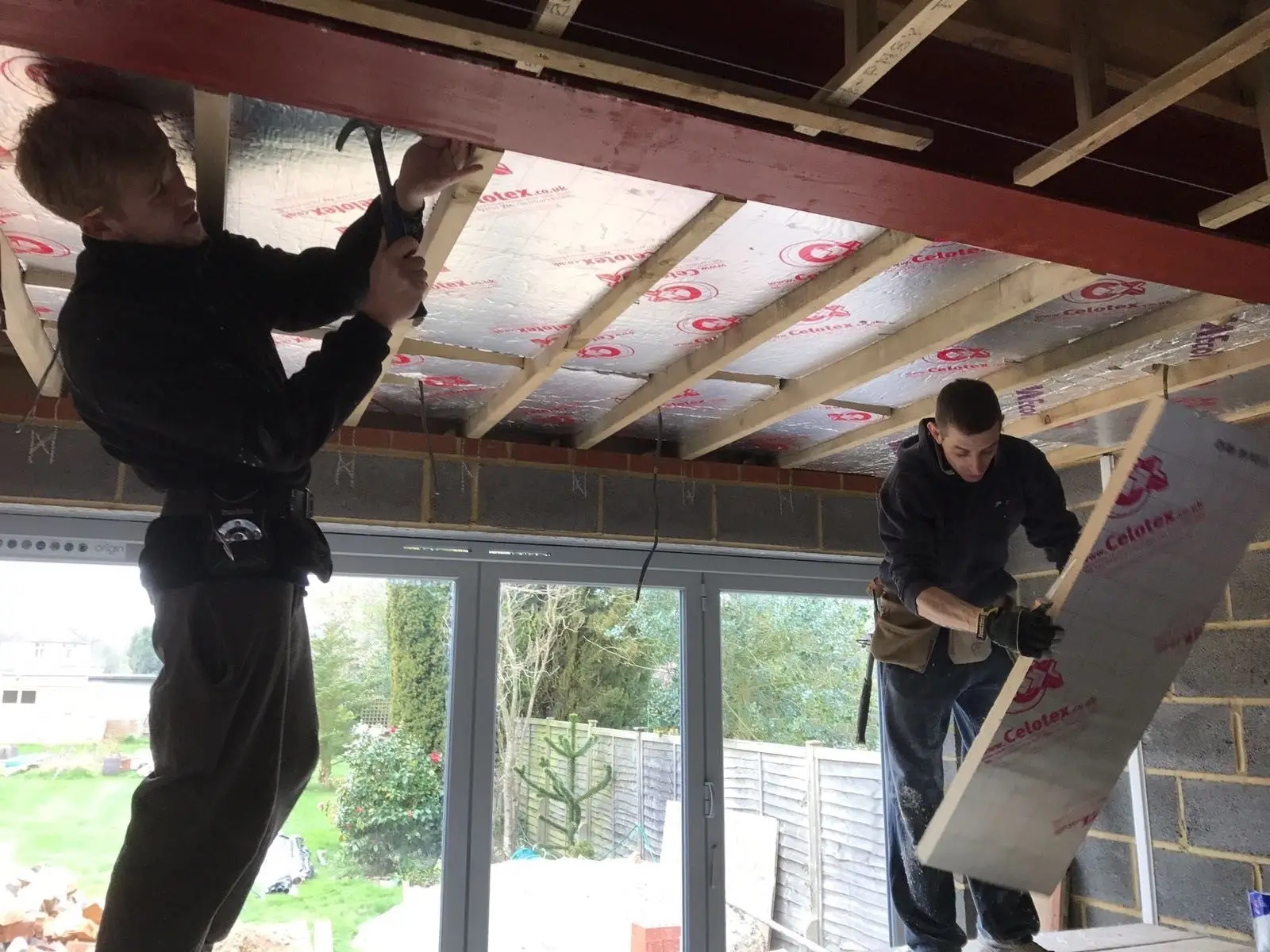 Builders pushing insulation boards up between new roof joists beside a red-painted steel beam