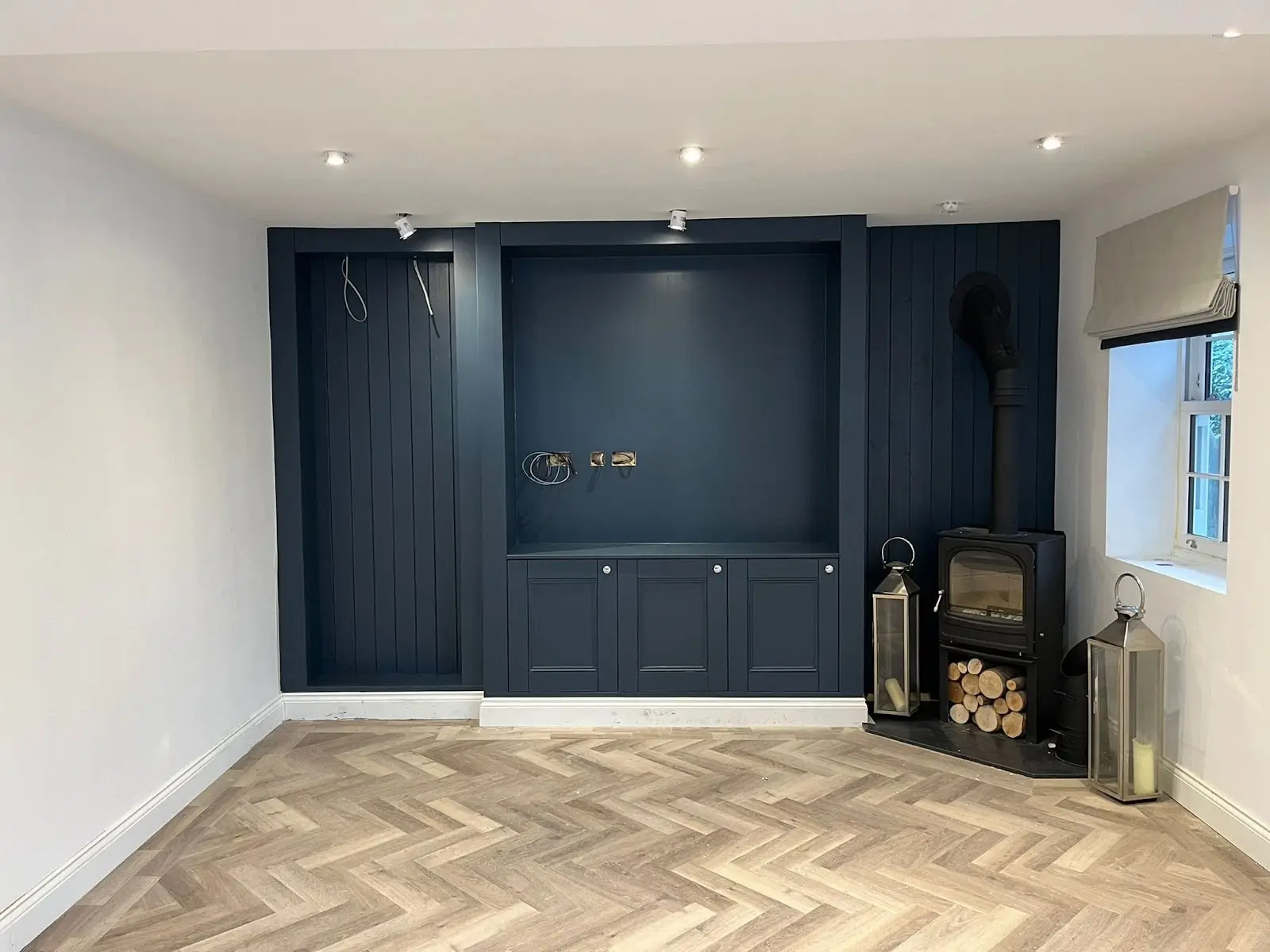 Navy fitted media wall with shelving, a log burner in the corner and herringbone flooring