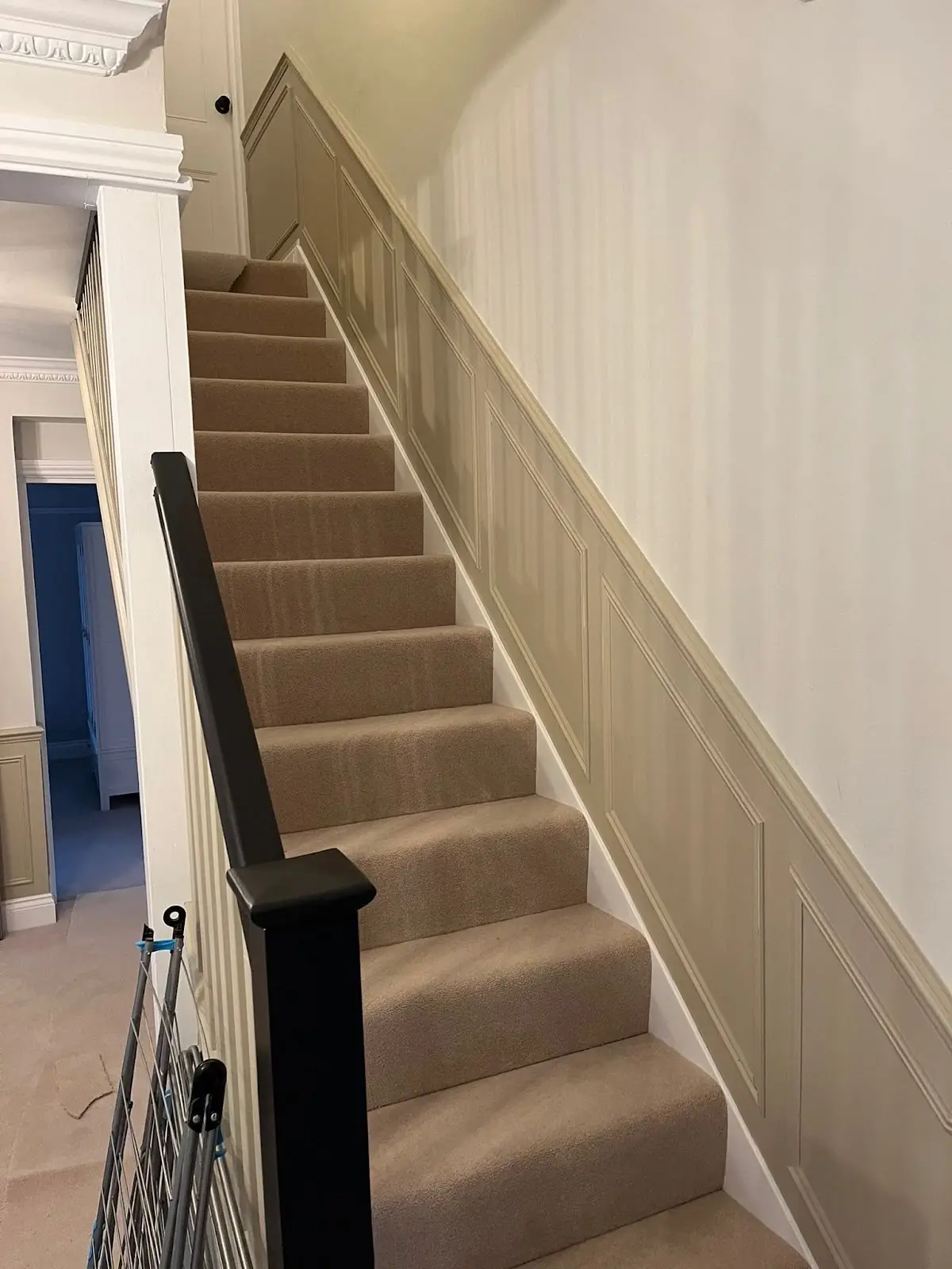 Stairs with a fitted carpet, painted panelling up the wall and a black handrail